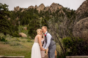 A bride and groom hold hands and smile at each other outdoors, with rocky hills and green trees in the background.