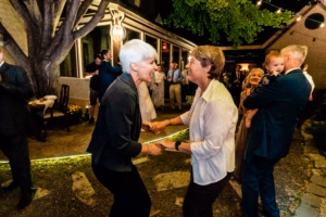 Two older women smile and hold hands while dancing outside at night, surrounded by other people at a social gathering.