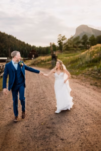 A bride in a white gown and a groom in a blue suit walk hand-in-hand down a dirt road surrounded by greenery and mountains.