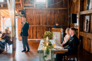 A man stands and speaks into a microphone at a Horstmann Homestead wedding reception as the bride and groom sit at a decorated table in a charming rustic wooden venue.