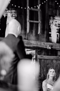 A woman in a light dress looks up at a man in a suit speaking; black-and-white photo with wedding portraits displayed on a rustic wooden wall at a Horstmann Homestead wedding.