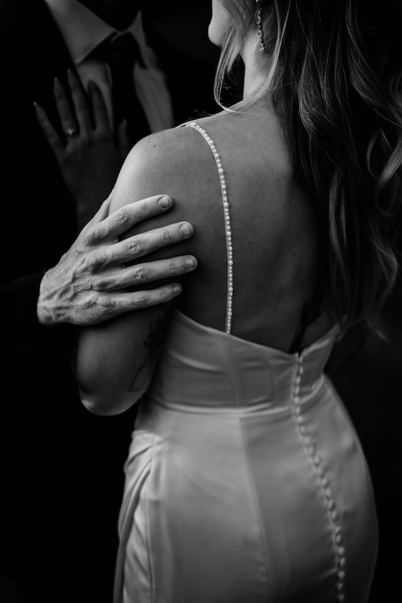 A man in a suit gently holds a woman in a white, buttoned dress from behind; the image is in black and white and focuses on their hands and upper bodies.