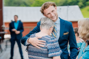 A young man in a blue suit smiles while hugging an older woman outdoors at a Horstmann Homestead wedding, with other people and a building visible in the background.