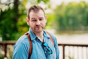 About us: Man with short hair and beard, wearing a blue striped shirt and sunglasses around his neck, stands outdoors near a wooden railing with trees and water in the background.