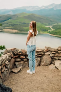 A woman in a white top and jeans stands on a stone lookout, overlooking a lake and green hills with mountains in the background, embodying the spirit of adventure that defines our About Us story.