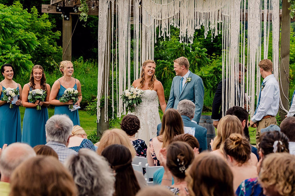 A bride and groom stand smiling at their Barn at Copper Creek Wedding, surrounded by bridesmaids in blue dresses, groomsmen, and guests seated outdoors during the ceremony.
