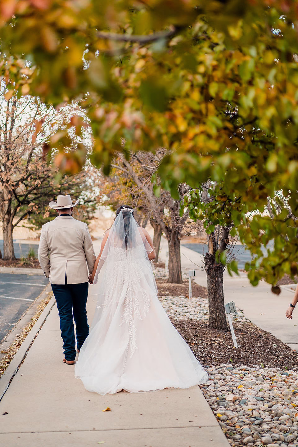 A bride in a white gown and veil walks hand-in-hand with a person in a tan jacket and cowboy hat along a tree-lined sidewalk.
