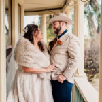 A bride in a lace gown and fur shawl stands beside a groom in a tan jacket and cowboy hat, smiling at each other on a porch.