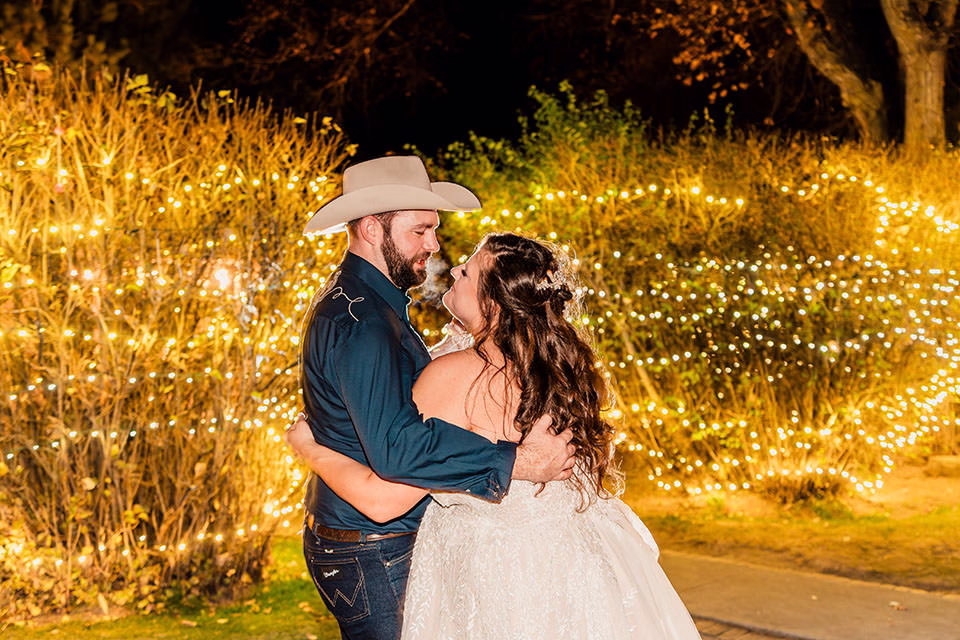 A bride and groom embrace outdoors at night, surrounded by string lights and illuminated foliage.
