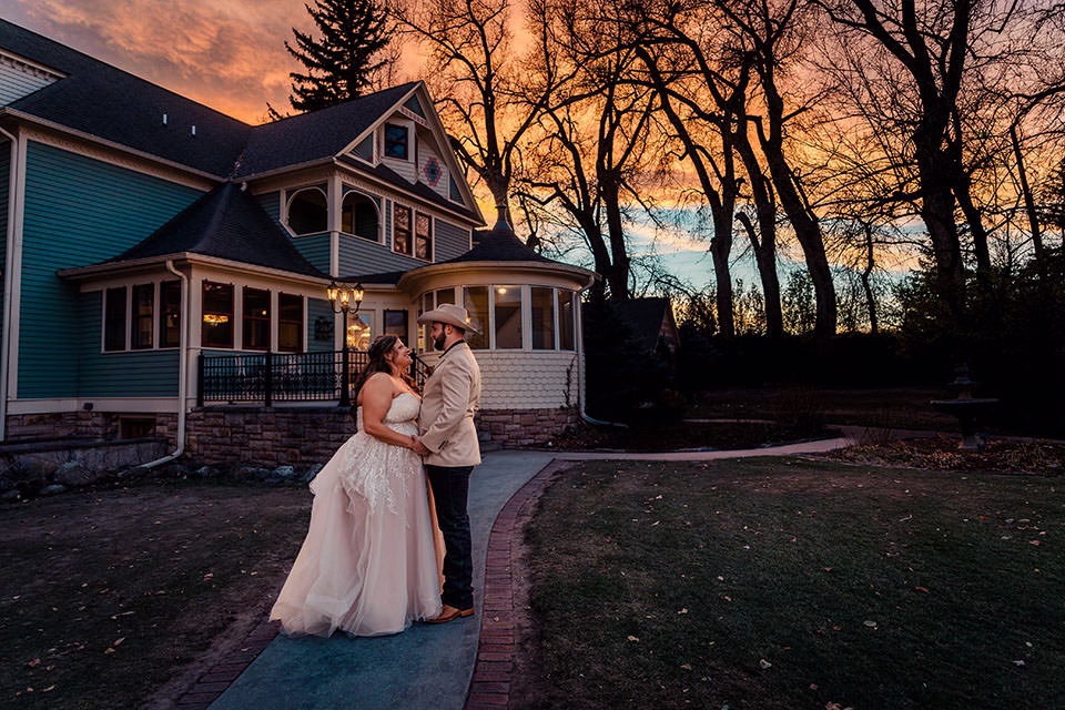A bride and groom stand facing each other outside a large house at sunset, with bare trees and a dramatic orange sky in the background.