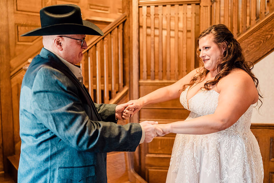 A man in a black hat and a woman in a white dress hold hands and face each other in front of a wooden staircase.