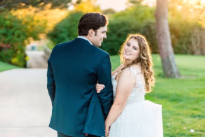 A bride and groom stand outdoors on a path at one of the stunning Colorado Wedding Venues, holding hands and facing each other, with greenery and sunlight in the background.
