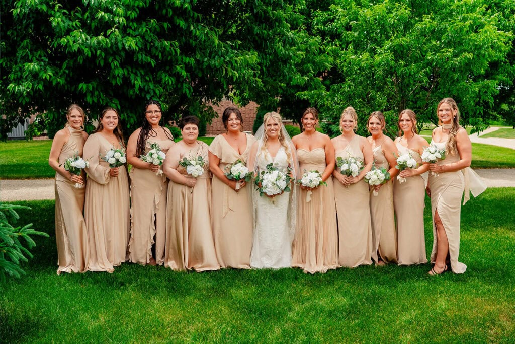 bridesmaids-in-champagne-dresses-outdoor-portraits-west-salem.jpg Wedding party of eleven women, including bride and bridesmaids, standing outdoors on a lush green lawn with trees in the background.