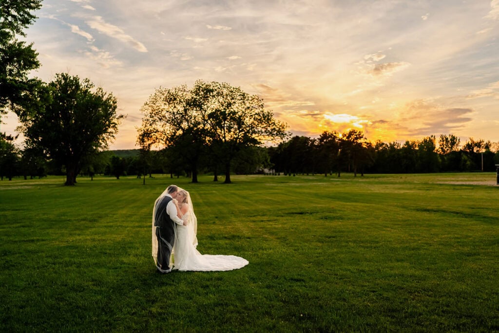 golden-hour-sunset-bride-groom-portraits-maple-grove-events-lawn.jpg Bride and groom sharing a kiss on a lush green field at sunset during a wedding at Maple Grove Events.