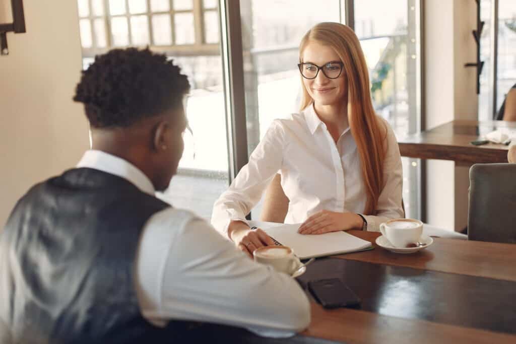 A Woman In A White Shirt And Glasses Smiling As She Interviews A Smartly Dressed Male Candidate Across A Table. There Is A Coffee Cup On The Table. White Text Overlay: '161 Job Interview Tips 2026: How To Excel In An Interview.'