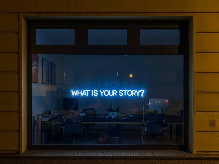 An image at brianvanderwaal.com/personal-profile-for-cv of a neon sign in a window which says "What is Your Story?" Through the window you can see an office with a desk, chairs, computers and other equipment. Your CV is a summary of your story, which is summarised in your personal profile even more succinctly.