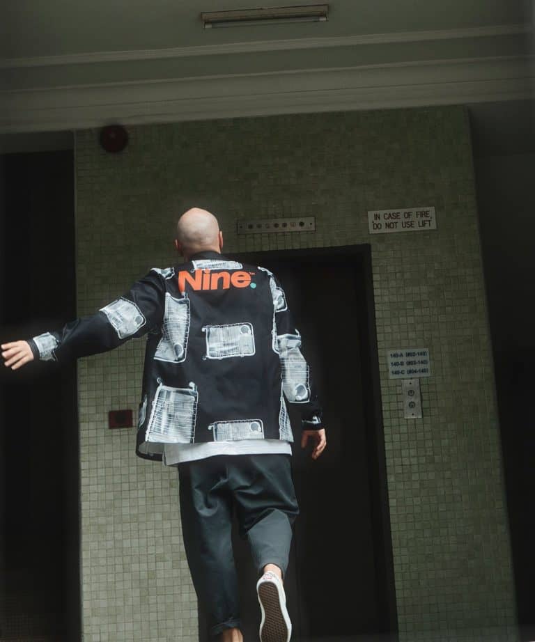 Man wearing a black streetwear jacket with white and red accents in an elevator lobby.