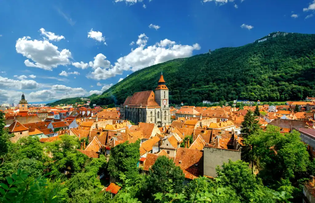 Historic Brașov town with red-tiled roofs and a prominent church, nestled beneath lush green mountai.