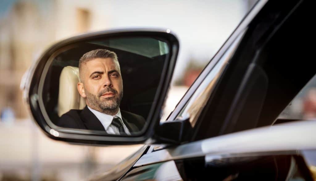 Professional chauffeur reflected in the side mirror of a premium vehicle