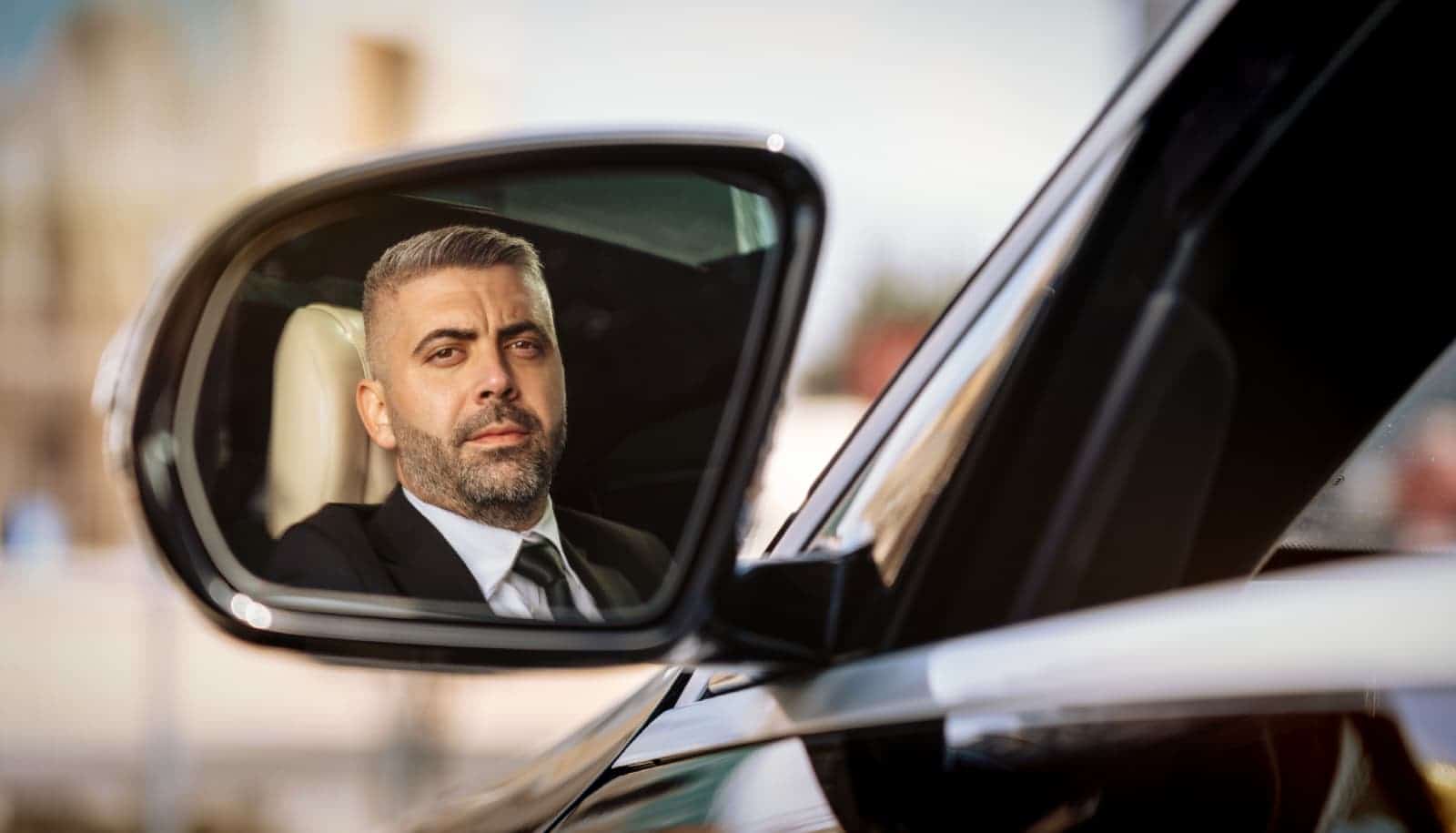 Professional chauffeur reflected in the side mirror of a premium vehicle