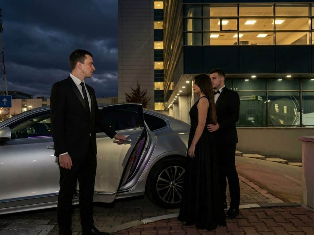 Chauffeur assisting elegantly dressed guests beside a premium sedan before an evening event
