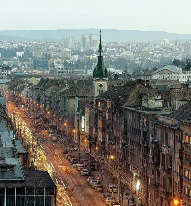 Urban street scene with old buildings, church spire, and city lights during evening hours.