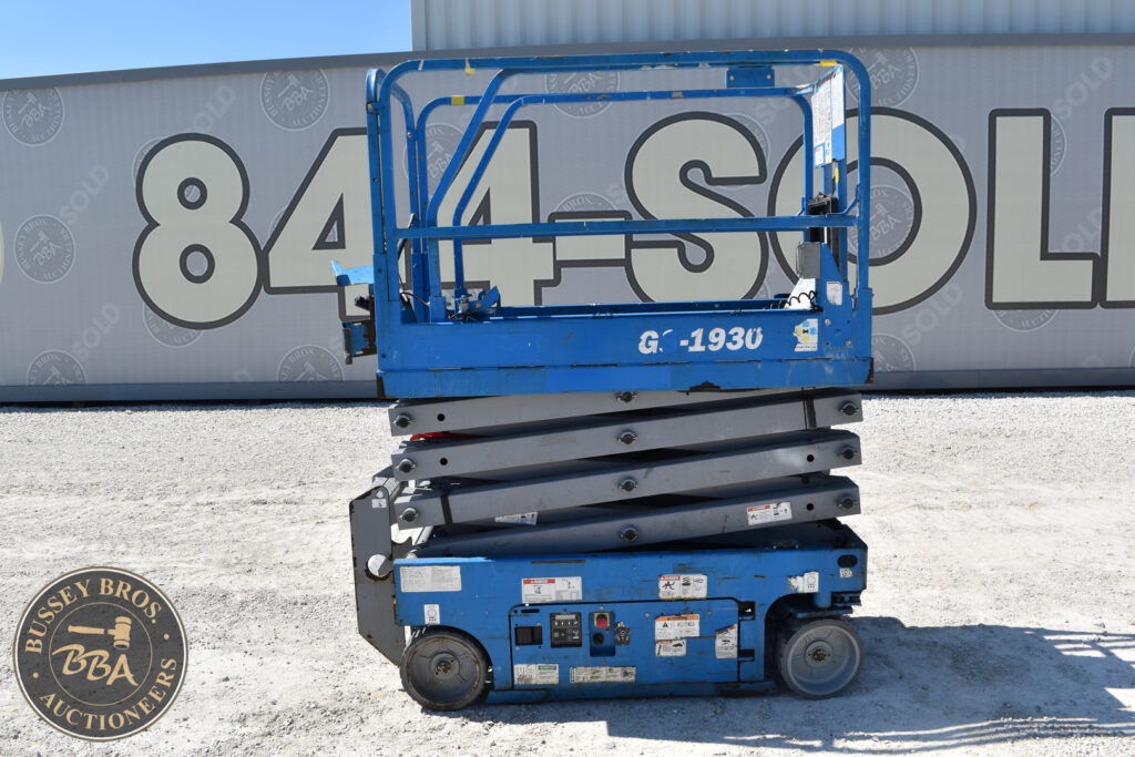 Blue scissor lift at auction site with large container in background.