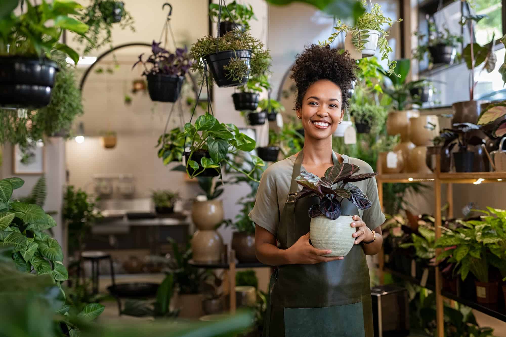 Vibrant young woman holding a potted plant in a lush indoor garden nursery, surrounded by hanging and shelf displays of various indoor plants, representing a plant shop or gardening store.