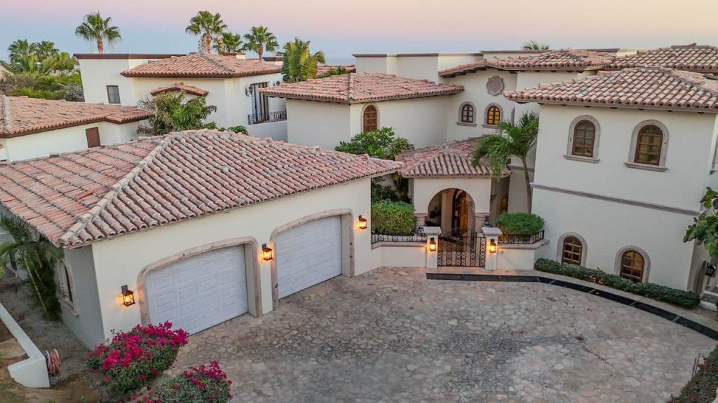 Elegant front exterior of Casa Alegria in Las Colinas, Cabo del Sol, showing the stone-paved driveway, double garage, arched entryway, and traditional Spanish-style architecture with tile roofs.