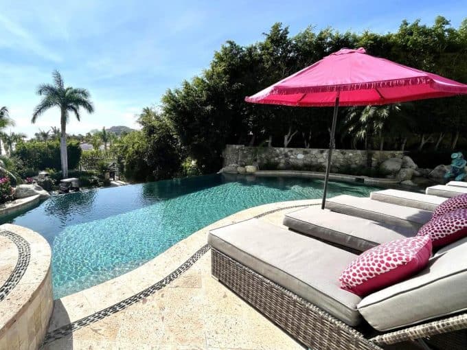 1. Luxurious Cabo Real Estate Pool With Pink Umbrella, Tropical Trees, And Scenic Ocean Views, Perfect For Relaxing.