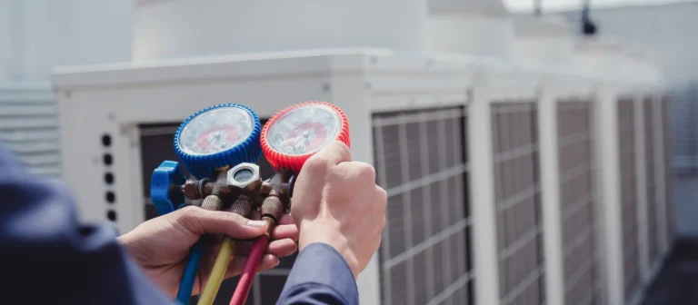 A person performs air conditioning maintenance by using a manifold gauge set to check the pressure of an HVAC system on a rooftop with several large air conditioning units.