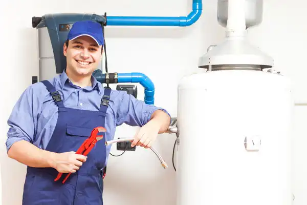 A plumber in blue overalls and cap stands next to a hot water heater, holding a pipe wrench and a flexible hose, ready for boiler maintenance and smiling at the camera.