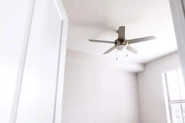 A white ceiling fan with five blades is mounted on a white ceiling in a brightly lit room, showcasing expert ceiling fan installation amid white walls and a window with blinds.