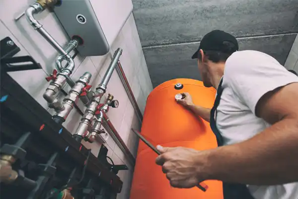 A worker in a cap inspects and adjusts controls on a large orange industrial tank surrounded by pipes in a utility room, ensuring efficient operation as part of general heating repair.