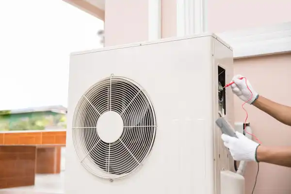 A person wearing white gloves uses a multimeter to check an outdoor air conditioning unit during routine heat pump maintenance.