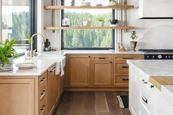 Modern kitchen with light wood cabinets, white countertops, open shelves with plants and dishes, a large window with forest view, farmhouse sink, and updated plumbing repair for peace of mind.
