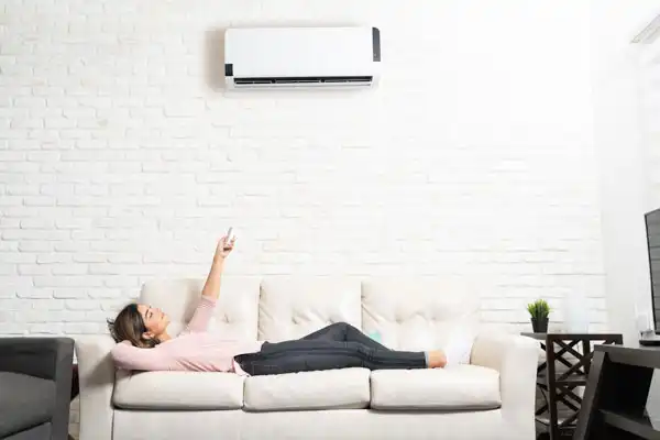 A person is lying on a sofa, pointing a remote control at a newly completed mini-split installation mounted on a white brick wall above them.