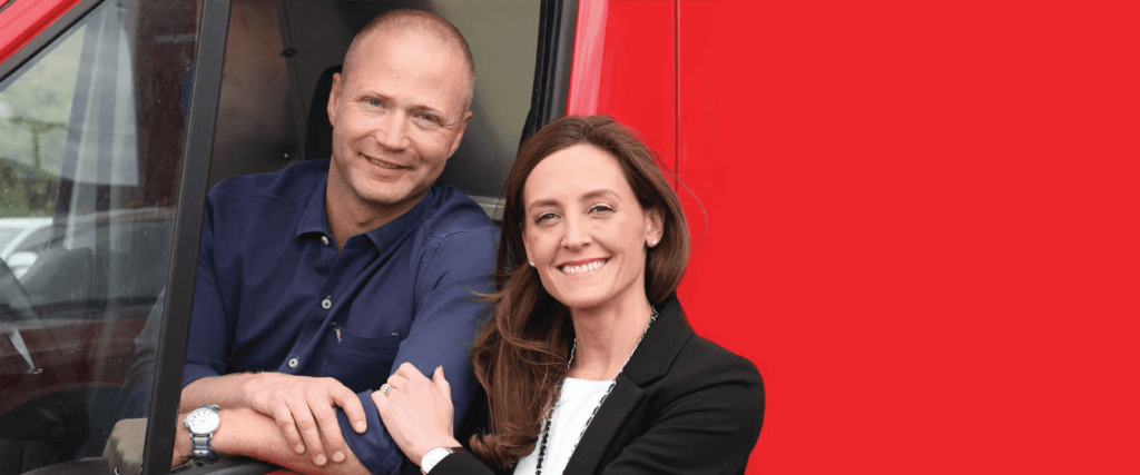 A man sits in the driver’s seat of a vehicle with the window open, while a woman stands next to him, both smiling at the camera in front of a red background.