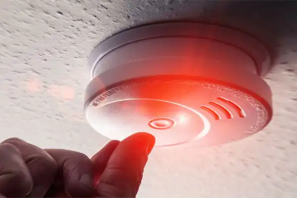 A person presses the test button on a ceiling-mounted Smoke And CO Detector, which is emitting a red light.