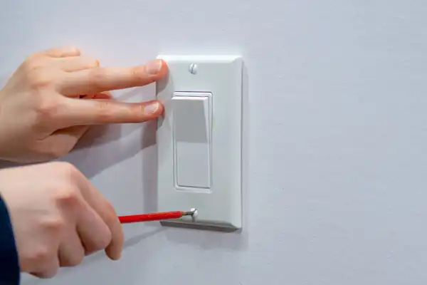 A person uses a screwdriver to tighten a screw on a white light switch plate, part of the electrical switches and outlets mounted on the wall.