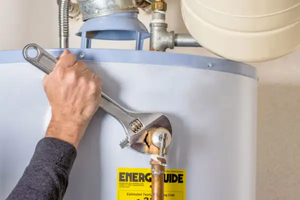A person uses a large adjustable wrench to tighten a pipe fitting on top of a water heater with an Energy Guide label, performing essential water heater repair.