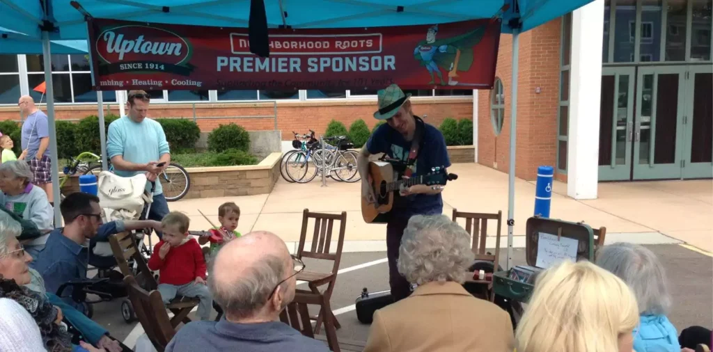 A musician plays guitar and sings under a blue tent to a seated audience at an outdoor event, with bicycles and sponsor banners visible in the background.