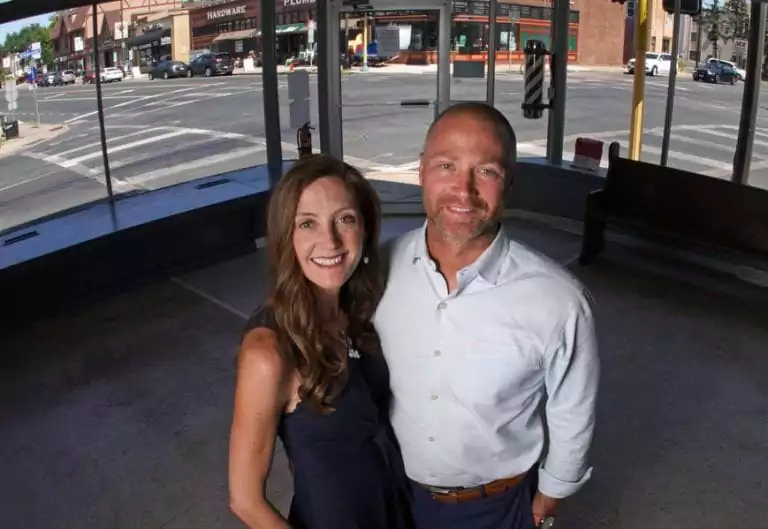 A woman and a man stand together inside a glass-walled building on a street corner, with shops and a crosswalk outside—capturing an everyday moment in the vibrant heart of the Twin Cities, as if waiting for their own hero news story to unfold.