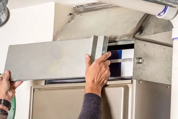 A person performs furnace maintenance by installing or removing a metal panel from an HVAC system, revealing the air filter inside.