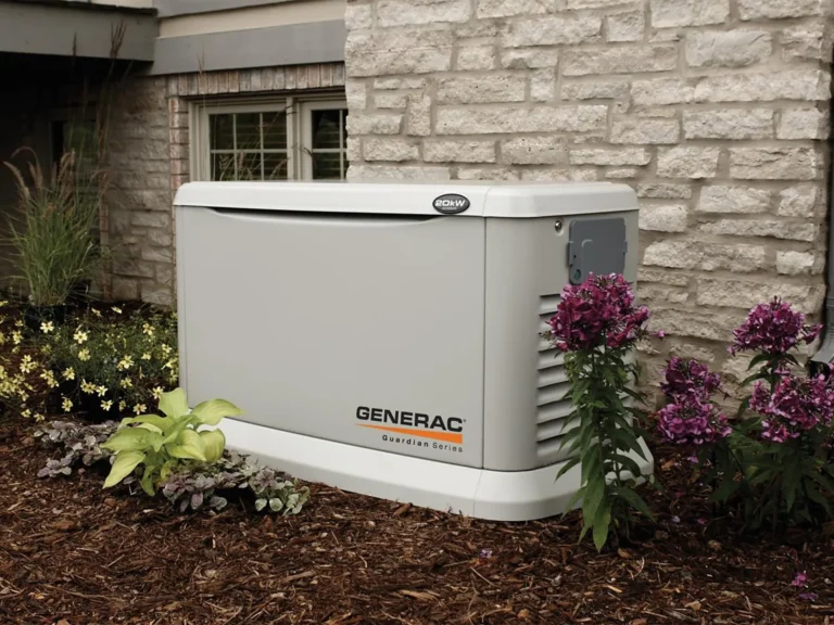 A Generac Guardian Series standby generator is installed outside a house as part of a professional home generator installation, surrounded by mulch, plants, and flowers, next to a brick wall and a window.
