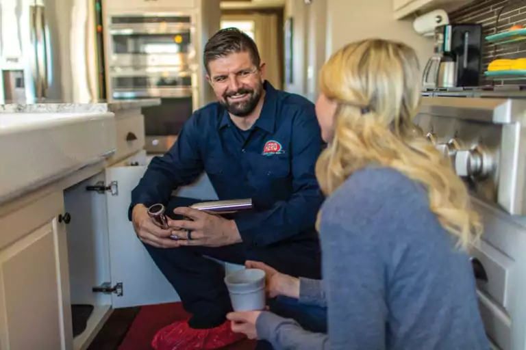A plumber sits on the kitchen floor near an open cabinet, talking to a woman holding a mug.