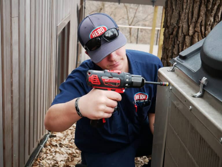 About Hero: A technician in a blue uniform and cap uses a red power drill to service an outdoor air conditioning unit next to a tree, showcasing the expertise and dedication that define a true hero in HVAC services.