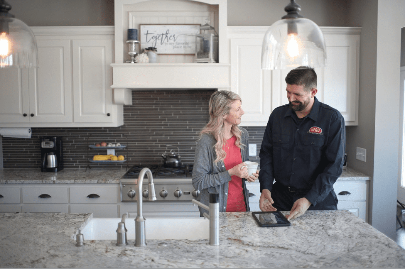 A woman and a man in a work uniform stand in a modern kitchen, discussing Twin Cities home services as they review information on a tablet on the counter.