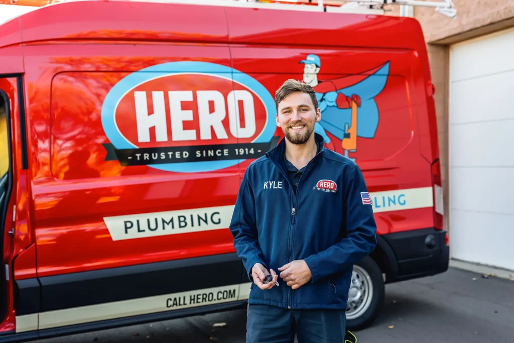 A man in a "HERO" uniform stands in front of a red HERO Plumbing van, smiling at the camera—showing what it’s all about Hero: friendly service and expert care.