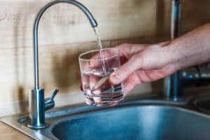 A person fills a clear glass with water from a kitchen faucet over a sink, focusing on water quality for a healthier New Year.